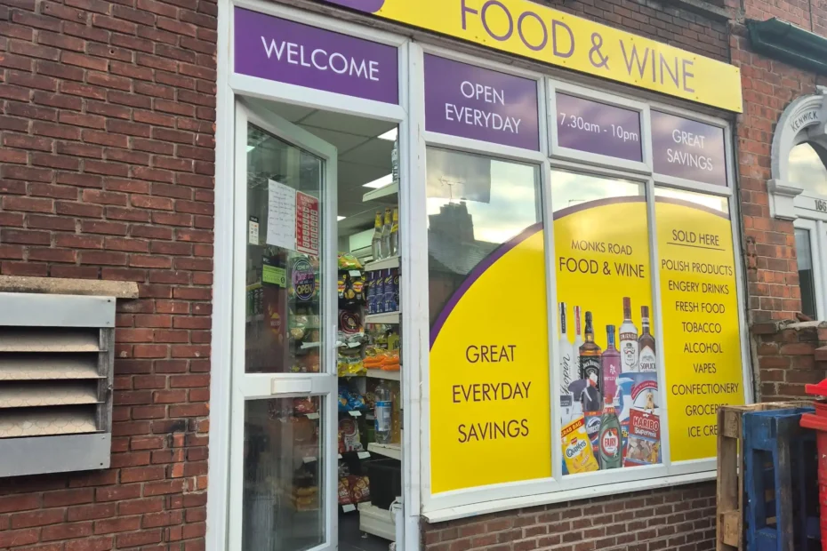 Storefront with purple and yellow signs advertising Food & Wine; brick building and glass door reveal grocery items inside, Monks Road branding visible.