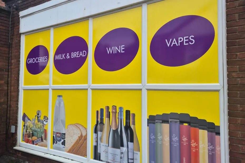 Storefront window with bright yellow panels and large purple circles listing product categories (groceries, milk & bread, wine, and vapes) with bottles visible at the bottom of the display window.