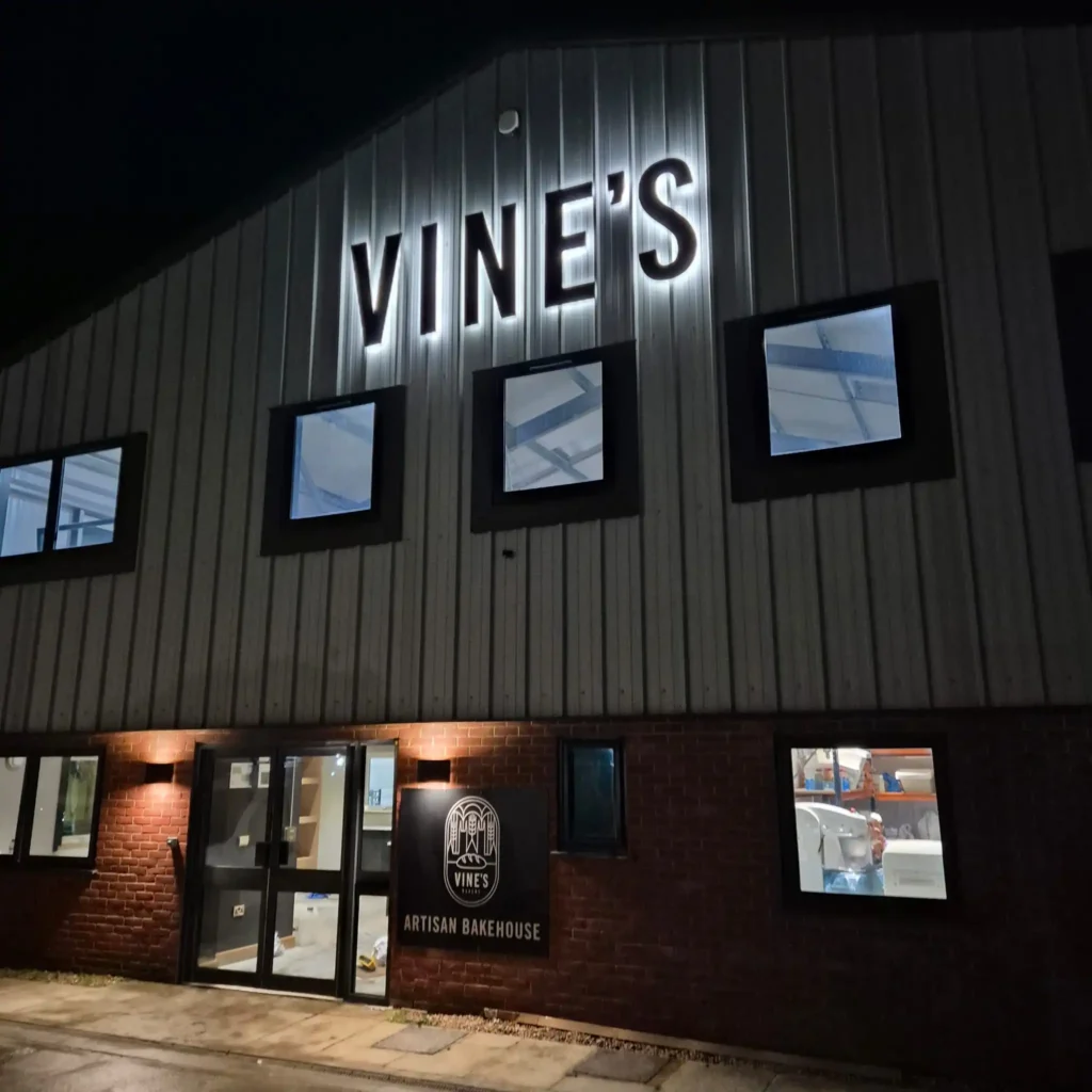 Night exterior of a brick bakery building with illuminated 'VINE'S' sign and 'ARTISAN BAKEHOUSE' logo near the entrance.