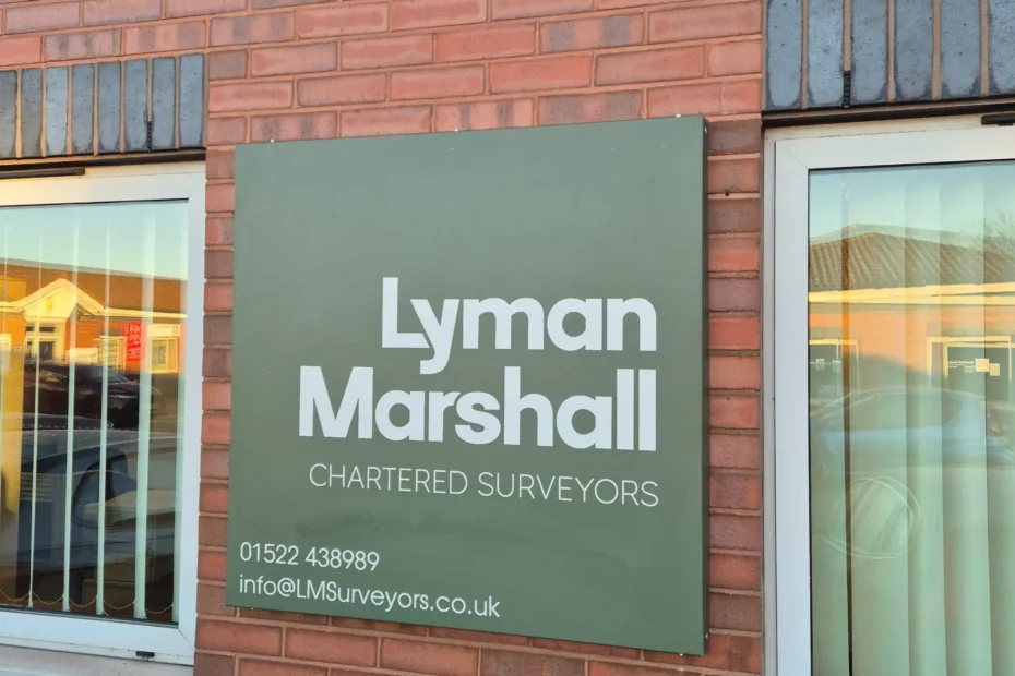 Brick exterior with a dark green sign displaying the business name (appears to read 'Lyman Marshall' Chartered Surveyors) and a blue 'Reserved Parking' sign below.