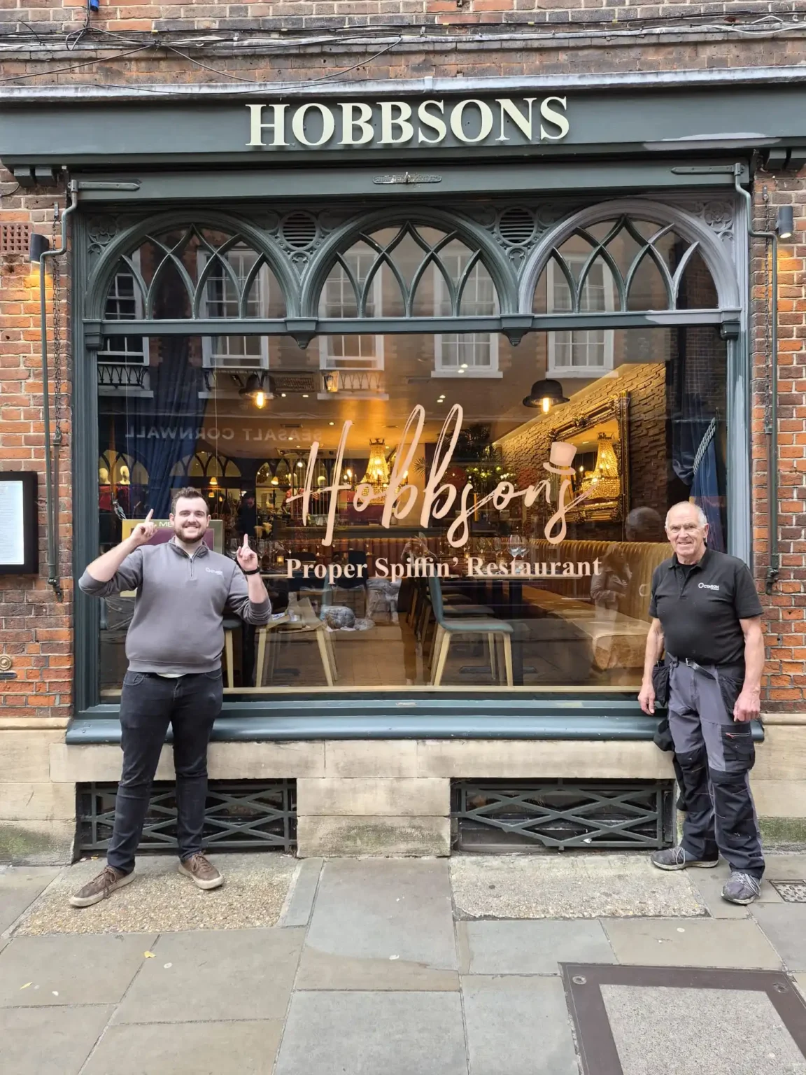 Two men stand outside a brick storefront with a large window for Hobbsons restaurant, smiling at the camera.