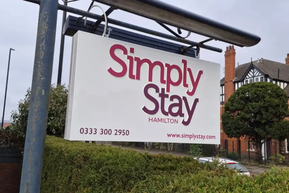 Sign reading 'Simply Stay Hamilton' with phone number 0333 300 2950 and website www.simplystay.com hanging above a hedge.