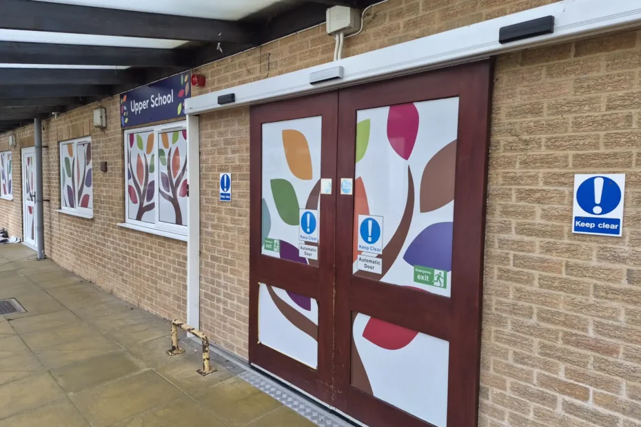 Brick school exterior with a purple double door covered in colorful leaf-pattern panels and an 'Upper School' sign above the doorway; several 'Keep Clear' and emergency exit signs are visible on the wall.