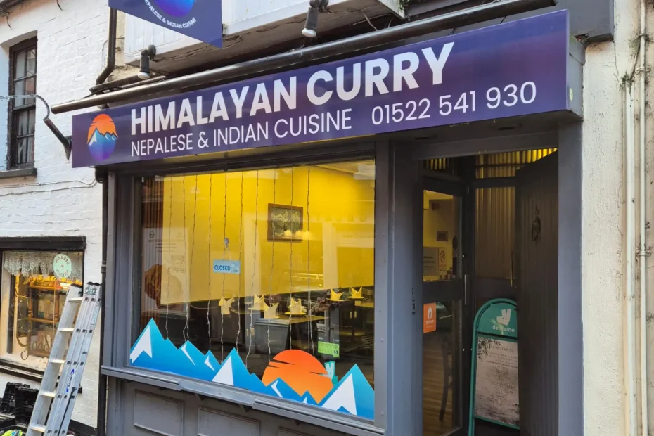 Storefront sign for Himalayan Curry Nepalese & Indian Cuisine, with a mountain logo and a ladder leaning against the window exterior.|Concise exterior view of Himalayan Curry restaurant sign with mountain graphic and purple-blue branding, ladder nearby|Shopfront advertising Himalayan Curry Nepalese & Indian Cuisine, mountain logo on blue sign above a large storefront window with a ladder beside it