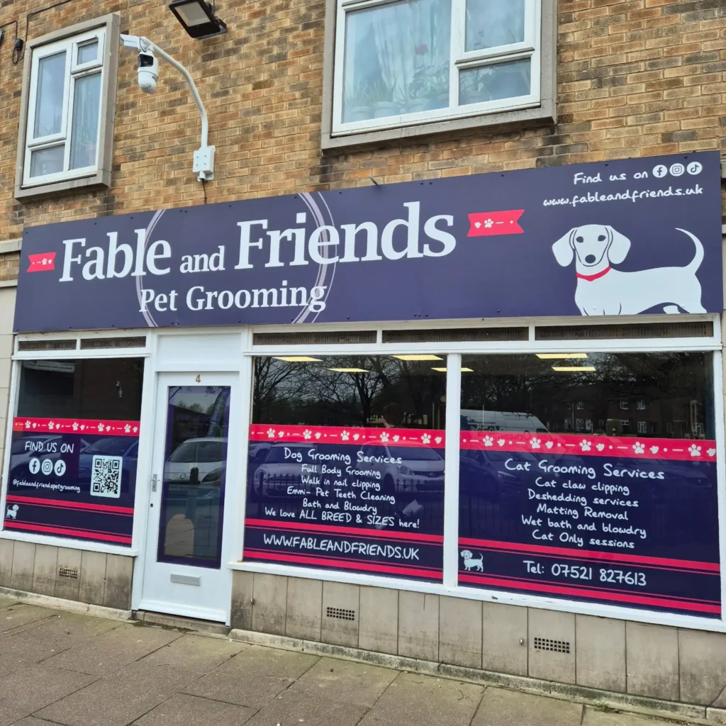 Storefront sign advertising 'Fable and Friends' pet grooming with a cartoon dog, brick building, and a security camera above.
