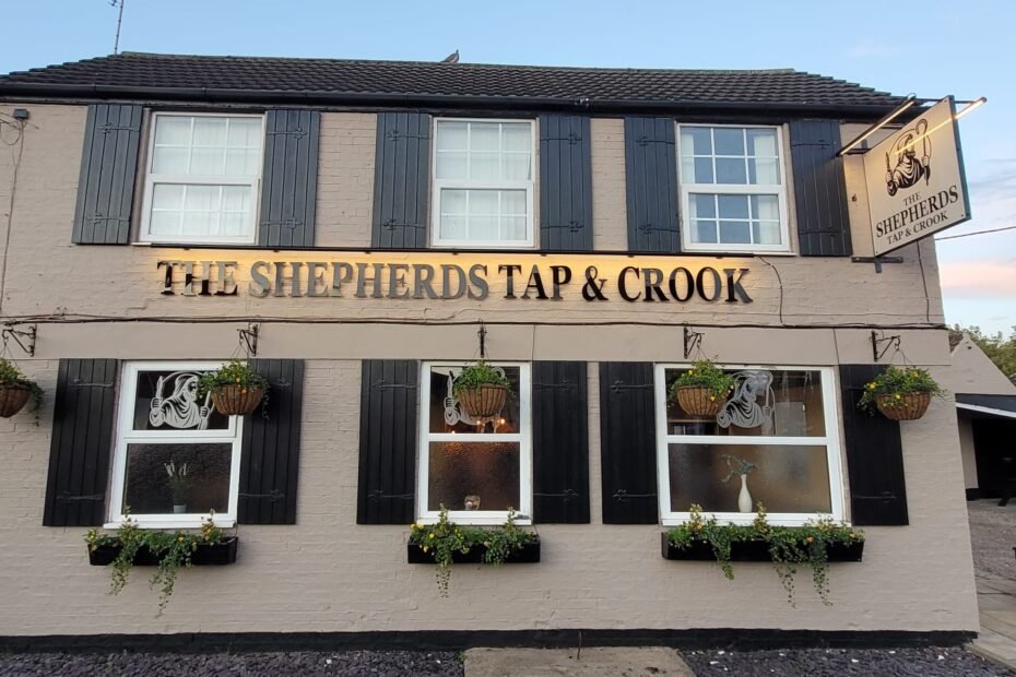 Front view of The Shepherds Tap & Crook pub: beige brick with black shutters, hanging baskets, and a large trough lighting bar above the entrance.