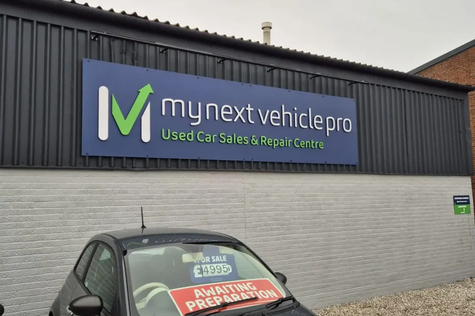 Front view of a gray car parked in front of a blue dealership sign reading 'my next vehicle pro – Used Car Sales & Repair Centre' on a dark corrugated metal wall.