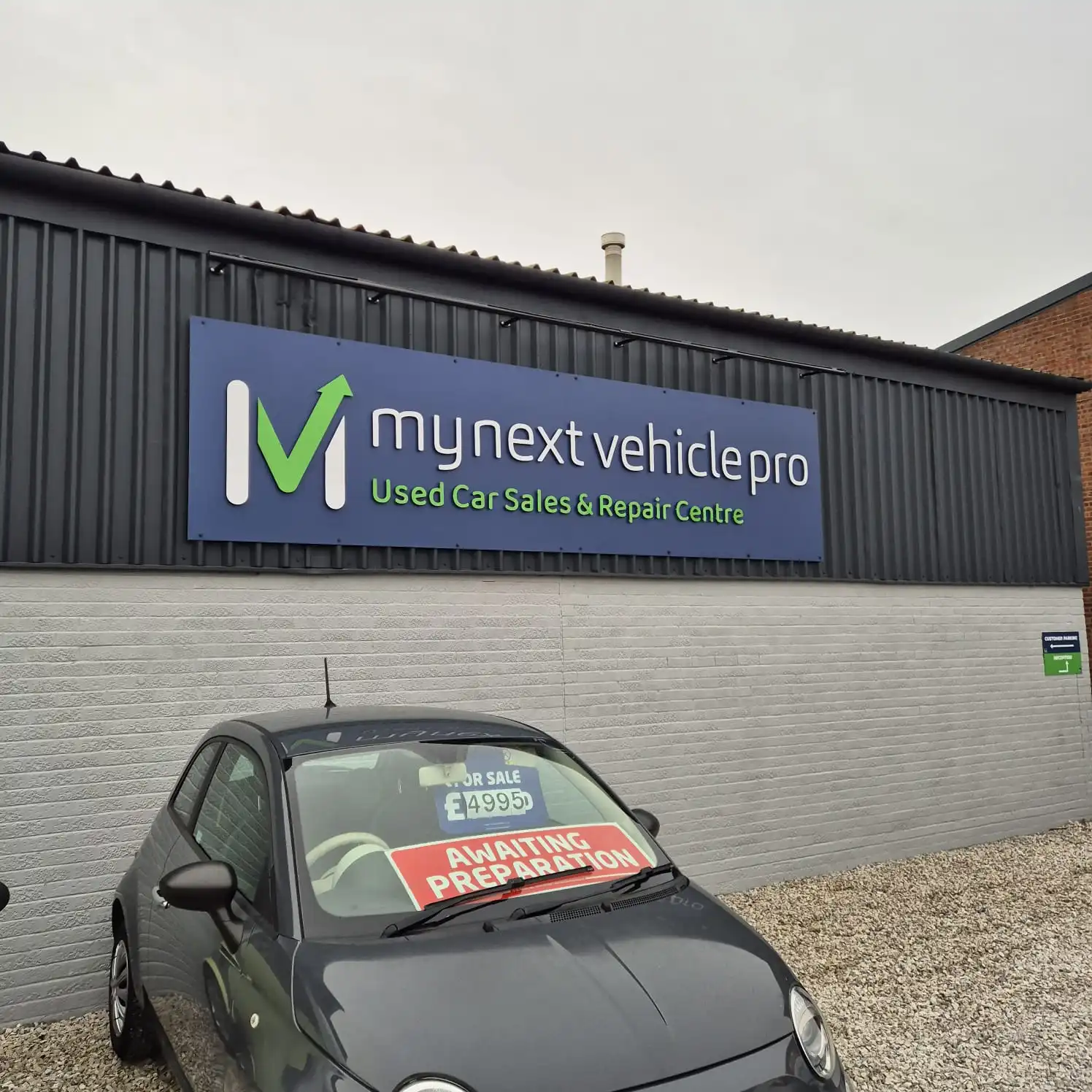Front view of a gray car parked in front of a blue dealership sign reading 'my next vehicle pro – Used Car Sales & Repair Centre' on a dark corrugated metal wall.