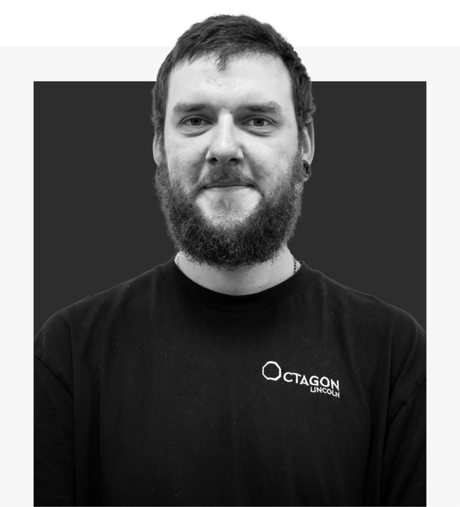 Close-up black-and-white portrait of a man with short hair and a full beard wearing a black shirt with an Octagon Lincoln logo.